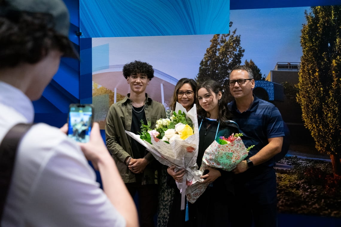 A person with a cell phone takes a photo of a grad with their family, holding flowers.