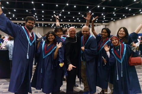Eight people wearing convocation robes raise their hands in the air in celebration.