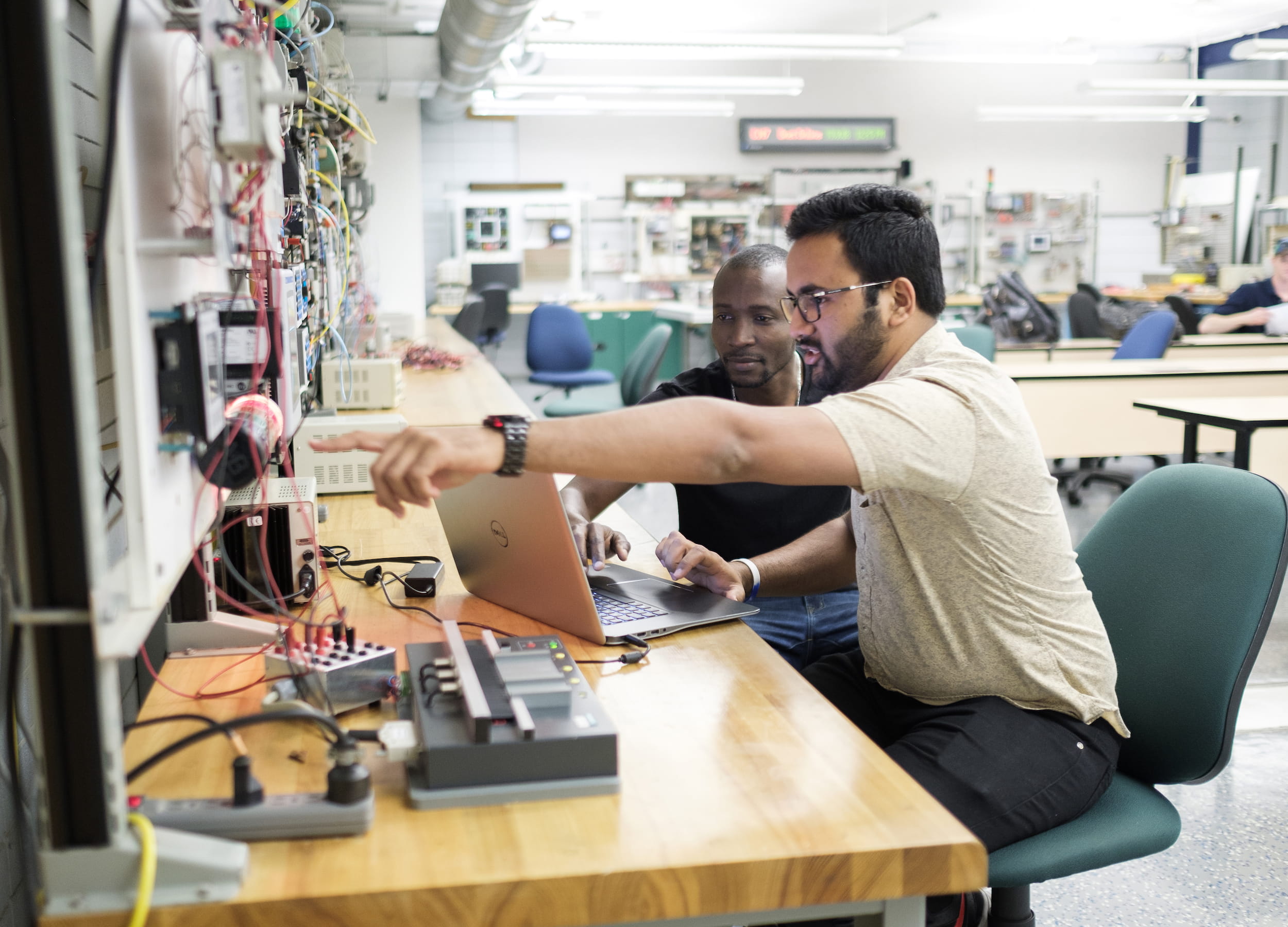 Two electrical engineering students sit on a laptop at a workstation. 